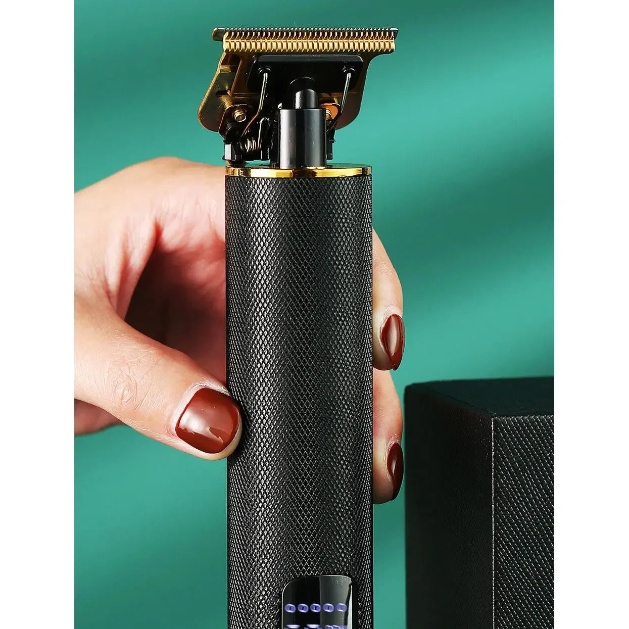 Hand holding black electric hair trimmer with gold blade on teal background